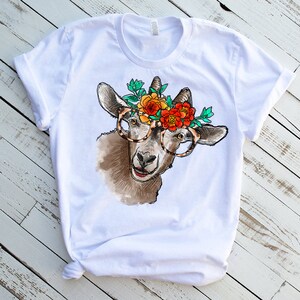 Op de afbeelding: Wit T-shirt met een afbeelding van een geit die een bril en een bloemenkrans draagt. De geit is bruin en wit met een zwarte neus en hoeven.