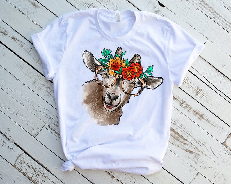 Op de afbeelding: Wit T-shirt met een afbeelding van een geit die een bril en een bloemenkrans draagt. De geit is bruin en wit met een zwarte neus en hoeven.