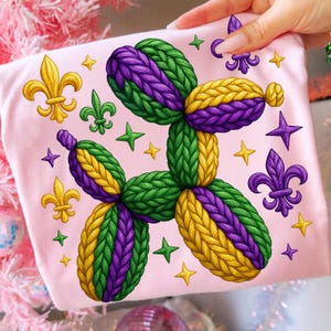 May include: A light pink t-shirt featuring a balloon dog design in green, purple, and yellow. The design is surrounded by gold fleur-de-lis, stars, and purple stars. The balloon dog is made to look like it is knitted.