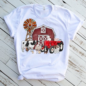 K&ouml;nnte beinhalten: Wei&szlig;es T-Shirt mit einem roten Stall, einer Windm&uuml;hle, einem roten Traktor und Nutztieren wie einer Kuh, einem Schaf, einem Schwein und einem Hund.