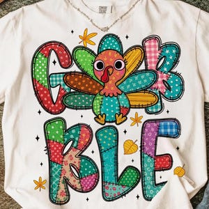 Könnte beinhalten: Weißes T-Shirt mit einem farbenfrohen Patchwork-Truthahn-Motiv und dem Wort "GOBBLE" im Patchwork-Stil. Der Truthahn hat einen roten Wamm und bunte Federn. Das Shirt hat ein Comfort Colors-Etikett.