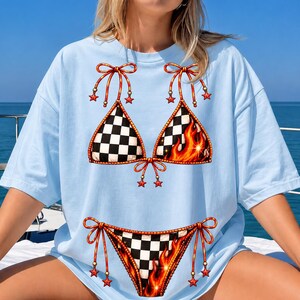 K&ouml;nnte beinhalten: Hellblaues &uuml;bergro&szlig;es T-Shirt mit einem Bikini-Motiv. Das Bikini-Oberteil hat auf einer Seite ein Schachbrettmuster und auf der anderen Flammen. Die Bikinihose hat ebenfalls ein Schachbrettmuster und Flammen. Rote Schleifen und Sternakzente vervollst&auml;ndigen das Design.