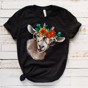 Op de afbeelding: Zwart T-shirt met een afbeelding van een geit die een bloemenkrans en een bril draagt. De geit kijkt naar rechts en heeft een vrolijke uitdrukking.