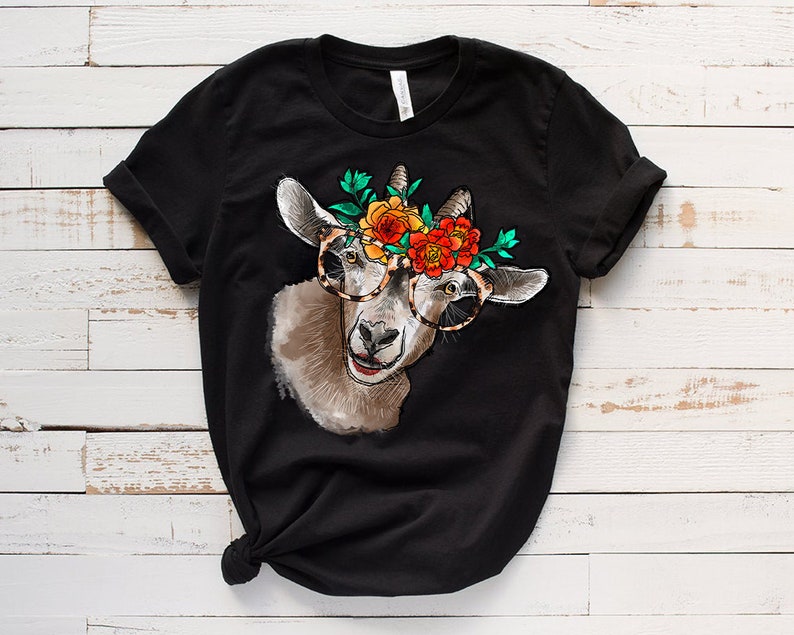Op de afbeelding: Zwart T-shirt met een afbeelding van een geit die een bloemenkrans en een bril draagt. De geit kijkt naar rechts en heeft een vrolijke uitdrukking.