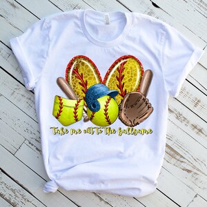 Puede incluir: Camiseta blanca con un dise&ntilde;o de tem&aacute;tica de softbol. El dise&ntilde;o presenta una pelota de softbol, un guante de receptor, un bate y un casco. El texto "Take me out to the ballgame" est&aacute; impreso debajo del dise&ntilde;o.
