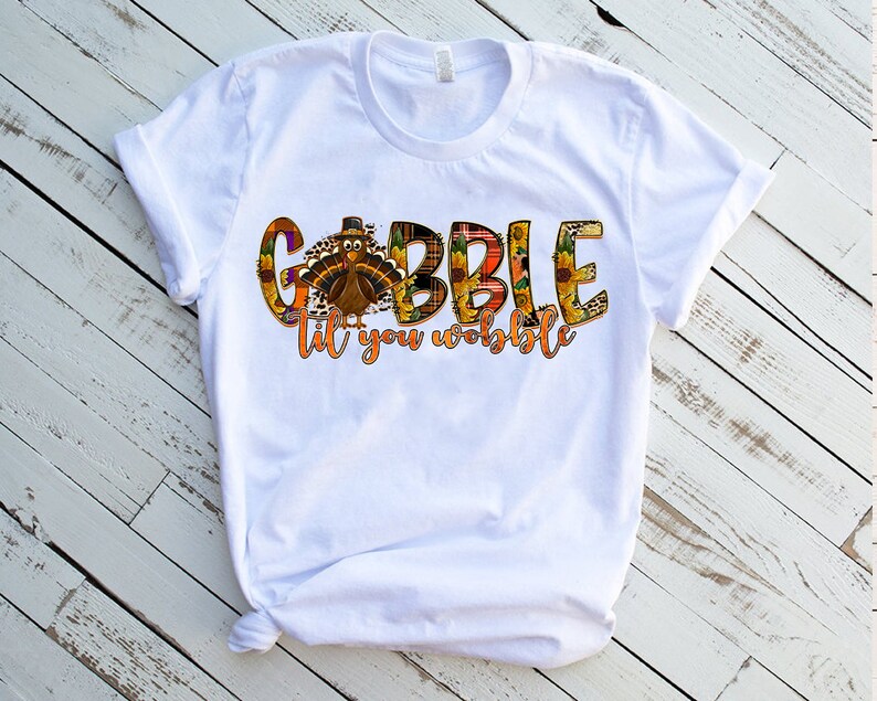 May include: White t-shirt with a Thanksgiving-themed graphic that says "Gobble til you wobble" with a turkey and fall-themed accents.