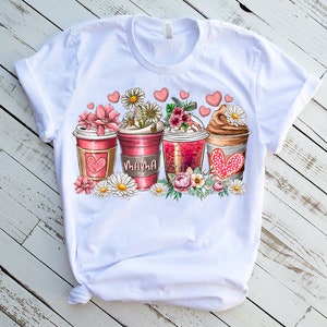 May include: White t-shirt with a graphic of three coffee cups with pink and red hearts, flowers, and the word "Mama" on one of the cups.