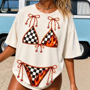 K&ouml;nnte beinhalten: Cremefarbenes T-Shirt mit Bikini-Design. Das Bikini-Oberteil und -Unterteil haben ein Schachbrettmuster und Flammen. Der Bikini ist mit roten Schleifen und sternf&ouml;rmigen Akzenten verziert. Das Shirt ist l&auml;ssige Sommerkleidung.