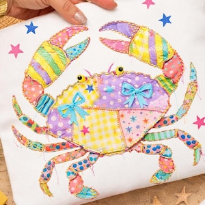 May include: A colorful crab applique on a white background, featuring patchwork patterns in various colors like yellow, purple, and pink. The crab has blue bows and is surrounded by star-shaped embellishments.