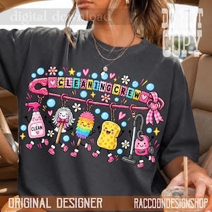 以下が含まれることがあります： チャコールグレーのTシャツで、CLEANING CREWという文字が入ったピンクの安全ピンの大きなグラフィックが描かれています。安全ピンからは、スプレーボトル、ダスター、スポンジ、掃除機など、顔のついた漫画風の掃除用具がぶら下がっています。