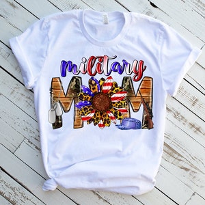 May include: White t-shirt featuring a sunflower graphic with red, white and blue accents, camouflage, and the text "military mom".