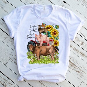 May include: White t-shirt with a graphic design of a pig, goat, cow, and birds on a red tractor. The text "Life is all about Balance" is printed on the shirt.