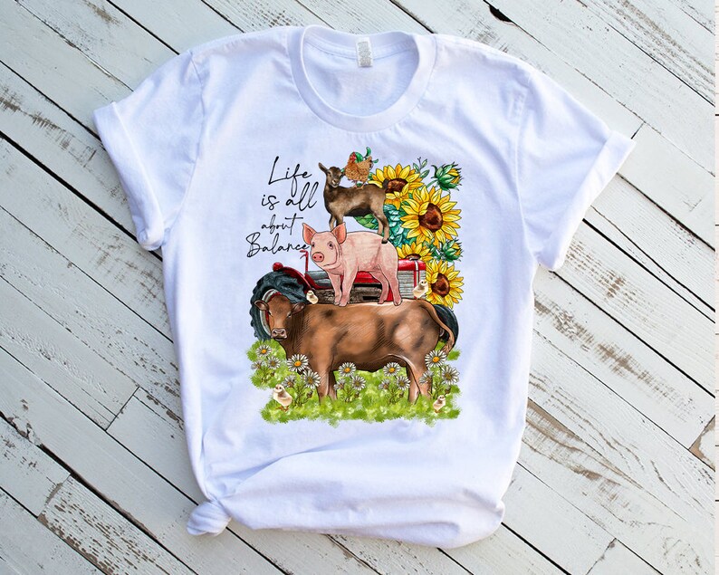 May include: White t-shirt with a graphic design of a pig, goat, cow, and birds on a red tractor. The text "Life is all about Balance" is printed on the shirt.
