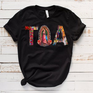 Puede incluir: Camiseta negra con la palabra "TOA" en letras de colores. La "O" es un c&iacute;rculo con una imagen religiosa en su interior.