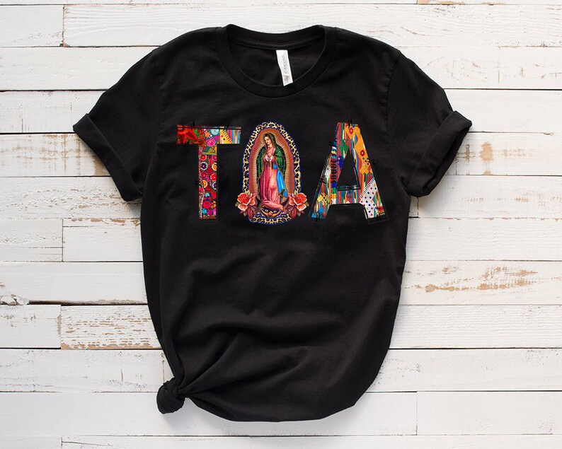 Puede incluir: Camiseta negra con la palabra "TOA" en letras de colores. La "O" es un c&iacute;rculo con una imagen religiosa en su interior.