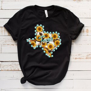 Puede incluir: Camiseta negra con un gr&aacute;fico del estado de Texas delineado con piedras de imitaci&oacute;n turquesa y relleno con un patr&oacute;n de piel de vaca y girasoles.