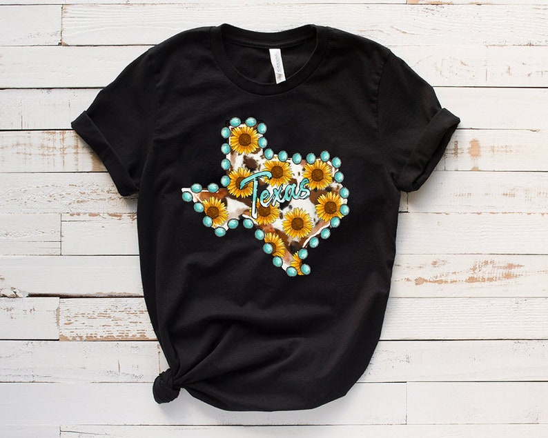 Puede incluir: Camiseta negra con un gr&aacute;fico del estado de Texas delineado con piedras de imitaci&oacute;n turquesa y relleno con un patr&oacute;n de piel de vaca y girasoles.