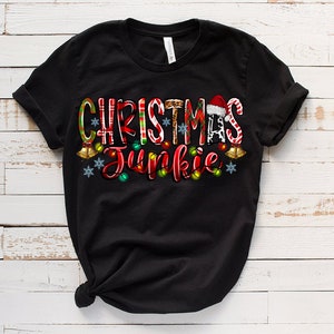 May include: Black t-shirt with the words "CHRISTMAS Junkie" in a festive design. The text incorporates Christmas elements like candy canes, a Santa hat, and string lights. The shirt is made of a soft material.