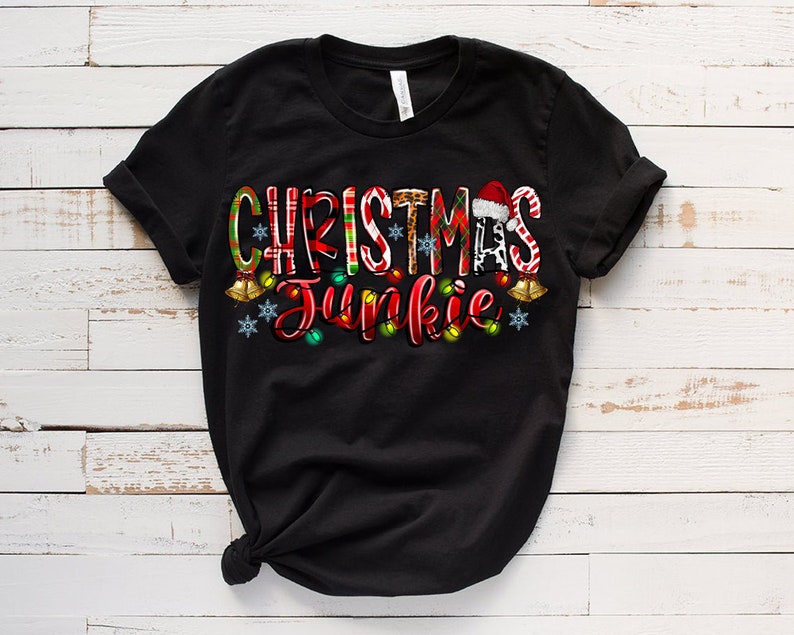 May include: Black t-shirt with the words "CHRISTMAS Junkie" in a festive design. The text incorporates Christmas elements like candy canes, a Santa hat, and string lights. The shirt is made of a soft material.
