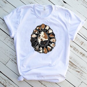 Pode incluir: Camiseta branca com um design estampado de concho prateado adornado, em couro de vaca marrom, preto e branco.
