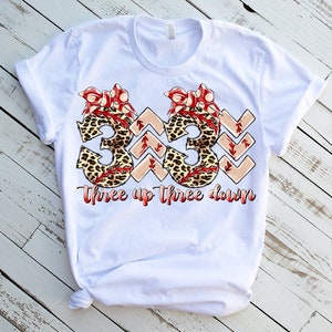 Puede incluir: Camiseta blanca con un gr&aacute;fico de dos n&uacute;meros 3 con estampado de leopardo y lazos rojos. El texto "three up three down" est&aacute; debajo de los n&uacute;meros.