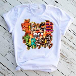 May include: White t-shirt with a colorful "Trick or Treat" graphic design. The design features a variety of patterns and textures, including stripes, leopard print, and animal print.