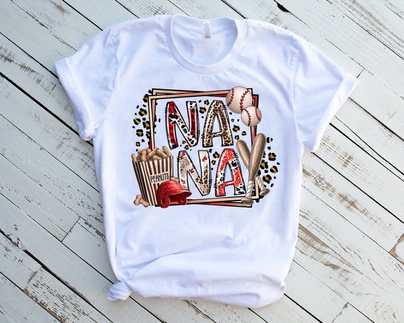 May include: White t-shirt with a graphic design featuring the word "NANAY" in red and black letters with a baseball theme. The design includes baseball bats, baseballs, peanuts, and a baseball cap.