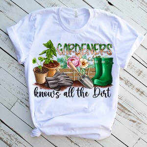 May include: White t-shirt featuring a green, brown, and pink gardening themed graphic. The graphic includes gardening gloves, a trowel, a watering can, and potted plants. The text on the shirt reads "Gardeners know's all the dirt".