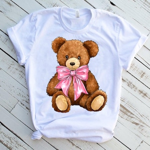 May include: White t-shirt with a brown teddy bear wearing a pink bow.