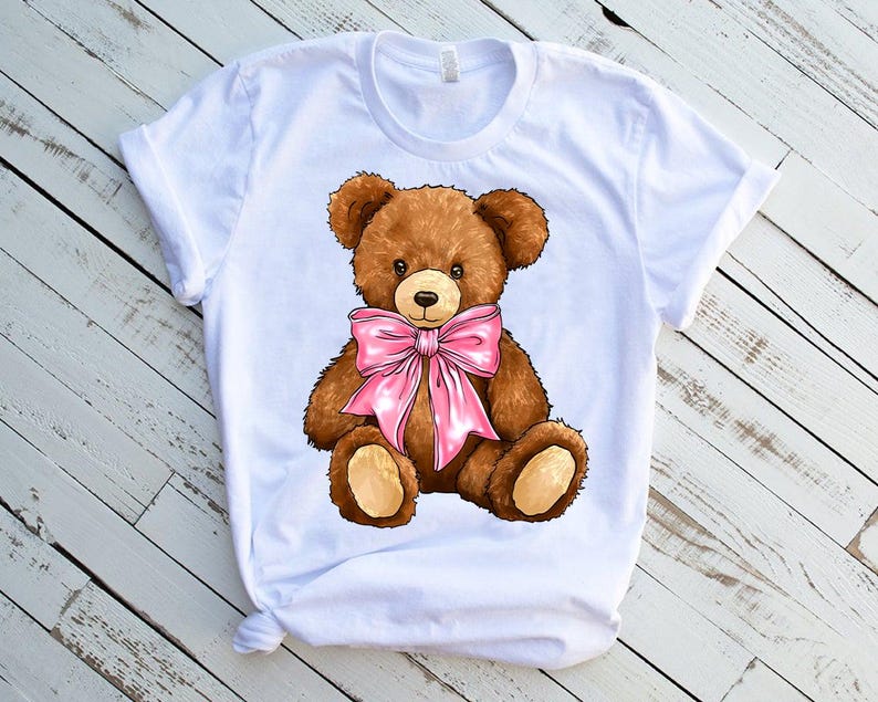 May include: White t-shirt with a brown teddy bear wearing a pink bow.