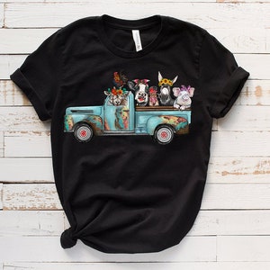 May include: A black t-shirt featuring a graphic of a blue pickup truck carrying a variety of farm animals, including a cow, pig, donkey, goat, and rooster. The animals are wearing flowers and are looking out of the truck bed.