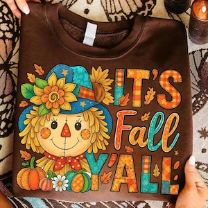 May include: Brown sweatshirt with a colorful graphic of a scarecrow wearing a blue hat with a sunflower and the text "IT'S Fall Y'ALL". The scarecrow has a red nose and a red bow tie. Pumpkins and fall leaves are also featured.