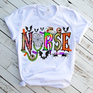 Puede incluir: Camiseta blanca con un dise&ntilde;o gr&aacute;fico colorido que dice "NURSE" con im&aacute;genes de tem&aacute;tica de Halloween como murci&eacute;lagos, ara&ntilde;as y una telara&ntilde;a.