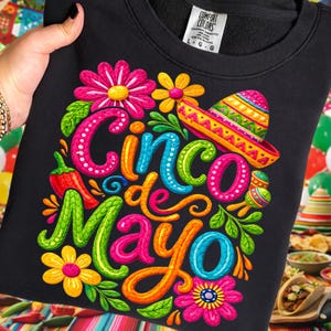 Peut inclure: Sweat-shirt noir avec un motif coloré du Cinco de Mayo. Le motif comprend le texte "Cinco de Mayo" aux couleurs vives, des fleurs, un sombrero et un piment.