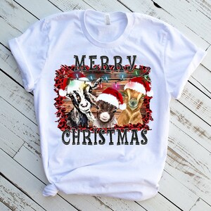 May include: White t-shirt with a Christmas design featuring three goats wearing Santa hats. The goats are in front of a wooden fence with Christmas lights. The text "Merry Christmas" is printed below the goats.
