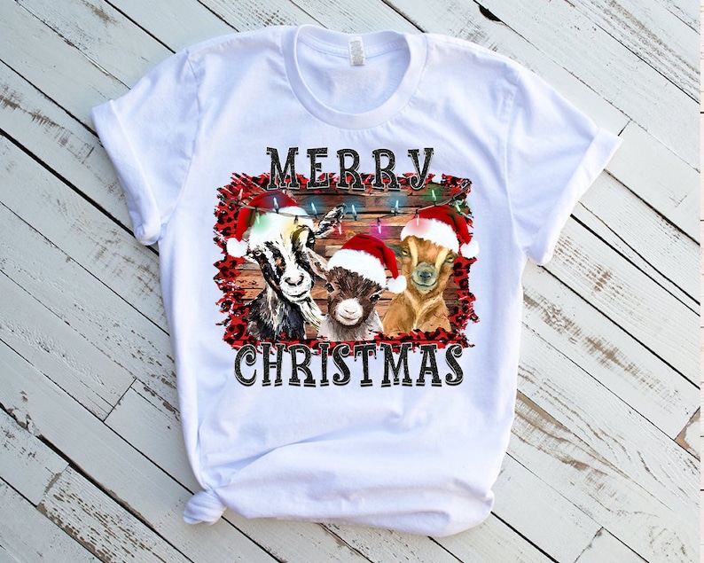 May include: White t-shirt with a Christmas design featuring three goats wearing Santa hats. The goats are in front of a wooden fence with Christmas lights. The text "Merry Christmas" is printed below the goats.