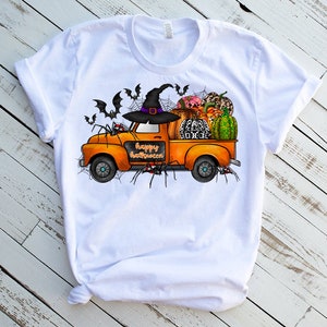 May include: White t-shirt with a graphic of an orange truck carrying pumpkins, gourds, and a witch's hat. The truck has the words "Happy Halloween" written on the side. There are also bats and spiders on the shirt.