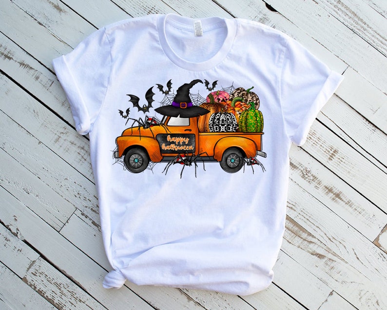 May include: White t-shirt with a graphic of an orange truck carrying pumpkins, gourds, and a witch's hat. The truck has the words "Happy Halloween" written on the side. There are also bats and spiders on the shirt.
