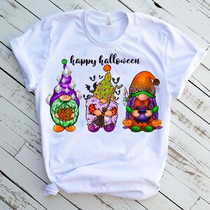 May include: White t-shirt with the text "happy halloween" and three gnomes wearing Halloween costumes. The gnome on the left is wearing a green and purple hat with white ghosts, the middle gnome is wearing a green and purple hat with an eye design, and the gnome on the right is wearing an orange and brown hat with purple hair.