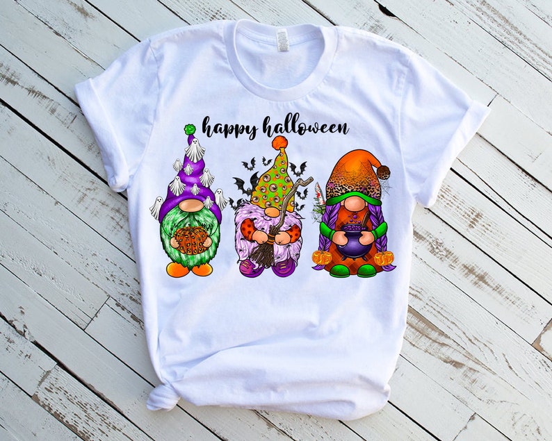 May include: White t-shirt with the text "happy halloween" and three gnomes wearing Halloween costumes. The gnome on the left is wearing a green and purple hat with white ghosts, the middle gnome is wearing a green and purple hat with an eye design, and the gnome on the right is wearing an orange and brown hat with purple hair.