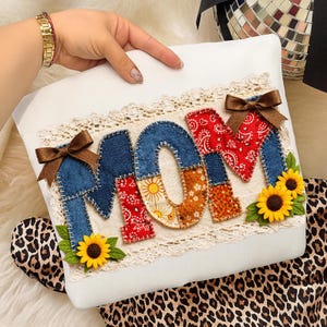 Può includere: Cuscino bianco con la scritta "MOM" applicata in denim, paisley e tessuti floreali. Le lettere sono delineate con un bordo cucito e decorate con fiocchi marroni e girasoli. Il cuscino ha un bordo in pizzo.