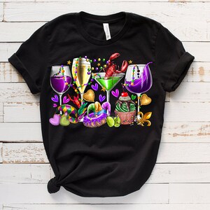 May include: Black t-shirt with a colorful Mardi Gras design featuring glasses of wine, a cocktail, beads, a cupcake, a donut, and a crawfish.