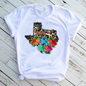 May include: White t-shirt with a graphic of the state of Texas in a leopard print with colorful flowers around it. The text "Texas Tough" is inside the state outline.