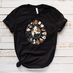Pode incluir: Camiseta preta com um design de estampa de couro de vaca marrom e branco no centro. O design &eacute; cercado por um c&iacute;rculo de acentos met&aacute;licos prateados.