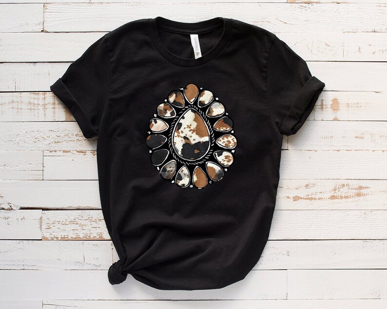 Pode incluir: Camiseta preta com um design de estampa de couro de vaca marrom e branco no centro. O design &eacute; cercado por um c&iacute;rculo de acentos met&aacute;licos prateados.