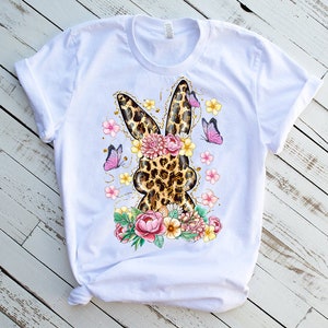 May include: White t-shirt featuring a leopard print bunny design with floral and butterfly accents. The bunny silhouette is surrounded by pink and yellow flowers and purple butterflies, creating a spring or Easter theme.