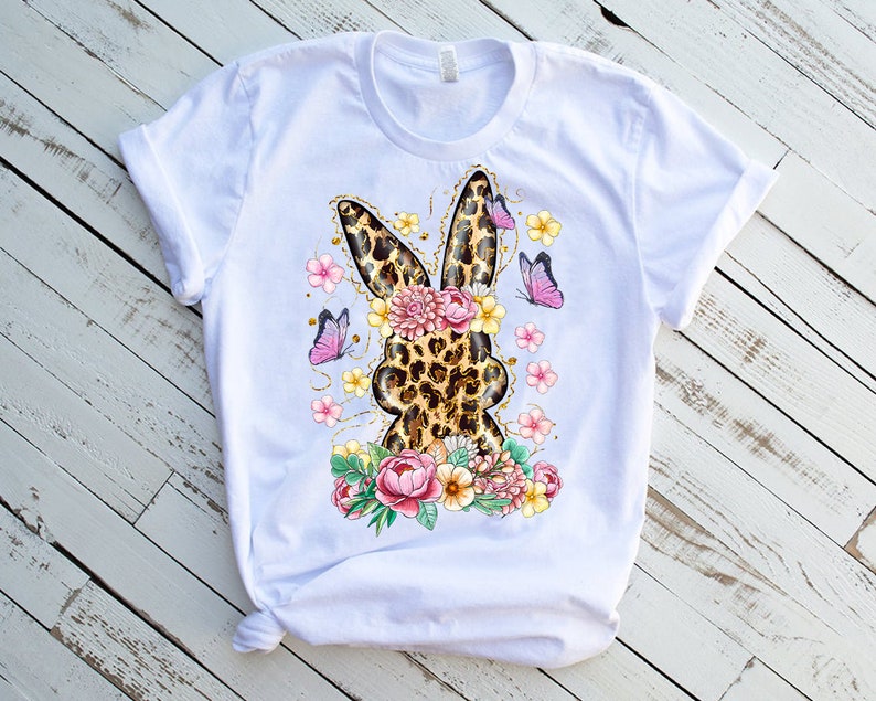 May include: White t-shirt featuring a leopard print bunny design with floral and butterfly accents. The bunny silhouette is surrounded by pink and yellow flowers and purple butterflies, creating a spring or Easter theme.