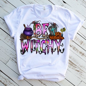 May include: White t-shirt with a pink and purple graphic that says "Be Witchy" with a cauldron, broom, bats, a tombstone, and a pumpkin.