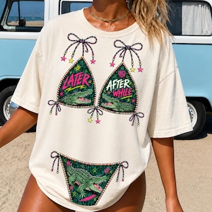 May include: Cream-colored t-shirt featuring a bikini design with the words "Later" and "After While" in neon green and pink. The bikini top and bottom have a green alligator graphic. The shirt has a relaxed fit.