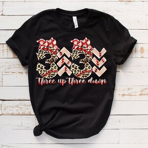 Puede incluir: Camiseta negra con un gr&aacute;fico de tres pelotas de b&eacute;isbol con estampado de leopardo y lazos rojos. El texto "Three up Three down" est&aacute; impreso debajo de las pelotas de b&eacute;isbol.
